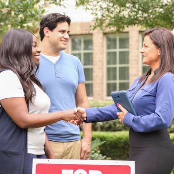 A young mixed-race couple shakes hands with a female real estate agent in front of a home that's for sale
