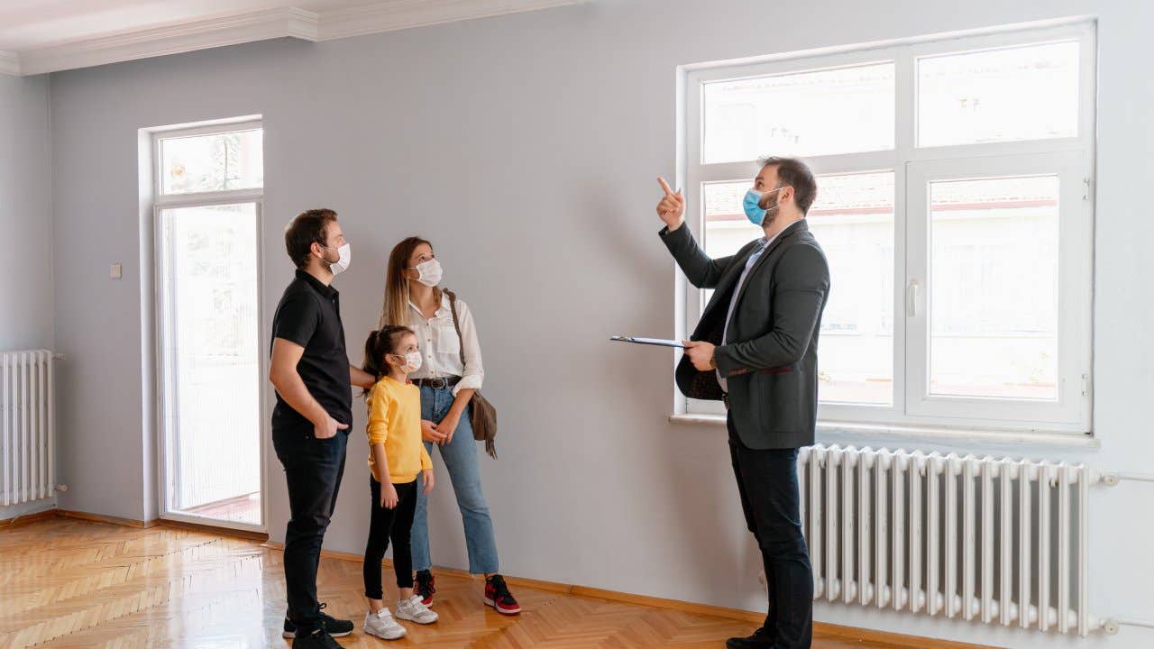 male real estate agent gestures toward a beautiful view out the window of a new home. Interested potential homeowners attentively listen to the real estate agent. All wearing surgical masks.