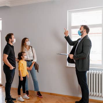 male real estate agent gestures toward a beautiful view out the window of a new home. Interested potential homeowners attentively listen to the real estate agent. All wearing surgical masks.