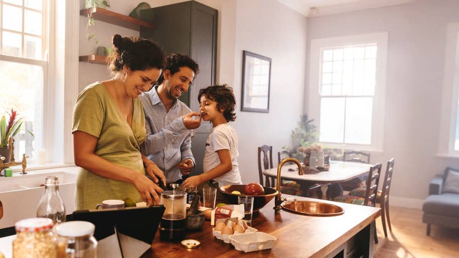 Playful dad feeding his son a slice of bread while his wife prepares breakfast.