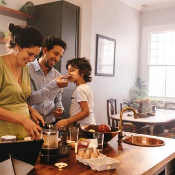 Playful dad feeding his son a slice of bread while his wife prepares breakfast.