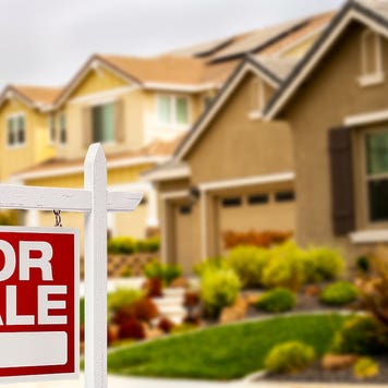 tan-colored suburban home with extensive landscaping and a red-and-white FOR SALE sign in front