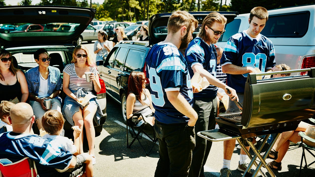 A group of adults on the younger side having a tailgate party in the parking lot full of cars. Some of them are wearing team jerseys and cooking on a grill while some others are holding drinks and sitting in the back of cars with the backdoors open.