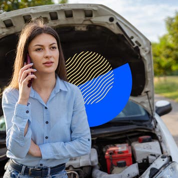 A driver standing in front of their car pulled off to the side of the road with the hood up; they are on their phone