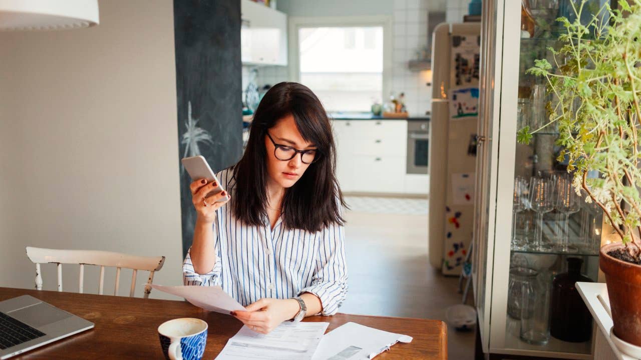 Woman with laptop and smartphone in dining room looking over bills