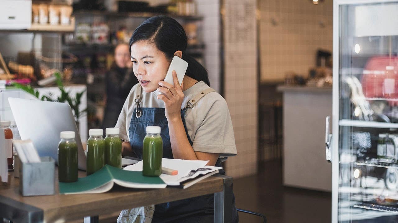 Asian female entrepreneur talking over smart phone while working on laptop in store