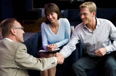 a seated couple holding paperwork smiling and shaking hands with a real estate agent