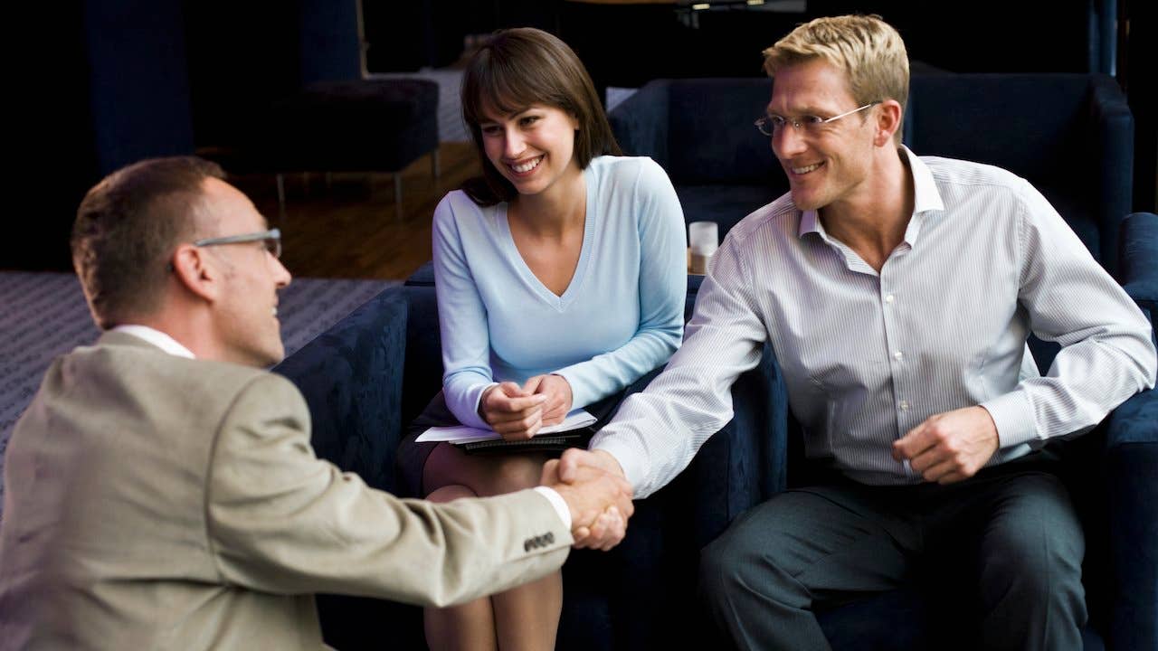 a seated couple holding paperwork smiling and shaking hands with a real estate agent