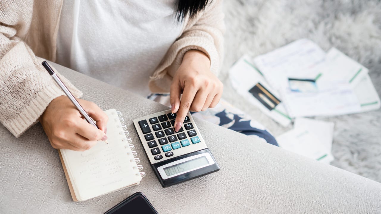 Woman writing a list of debt on notebook calculating her expenses with calculator with many invoices