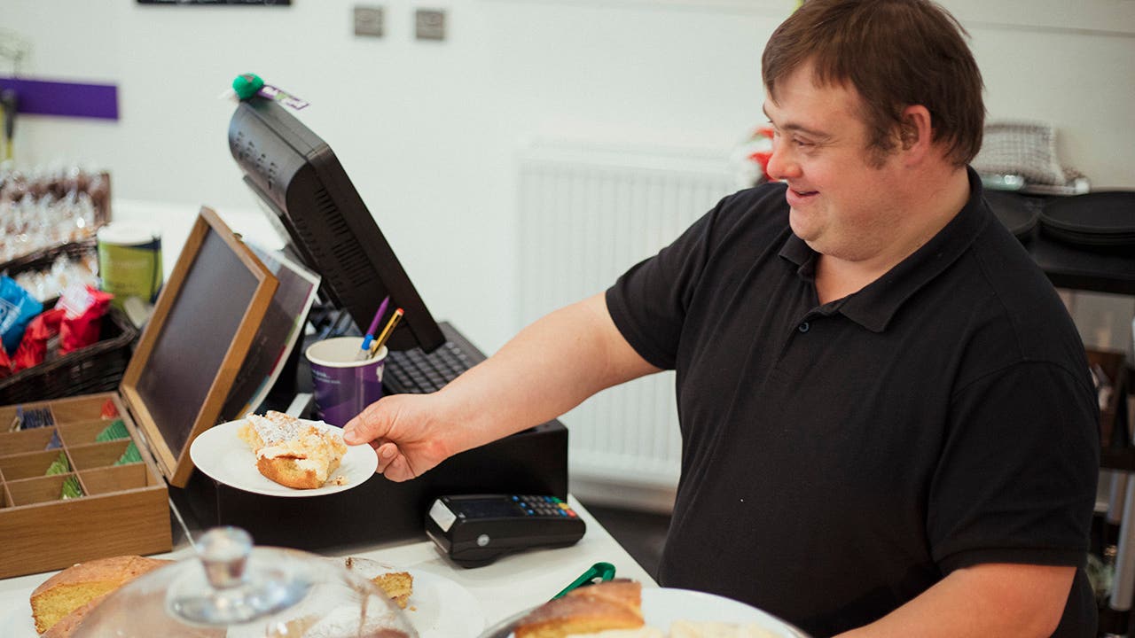 A man with Down syndrome is serving cake in a cafe where he works.