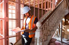 A builder sits on the stairs inside a house under renovation and studies the building plans