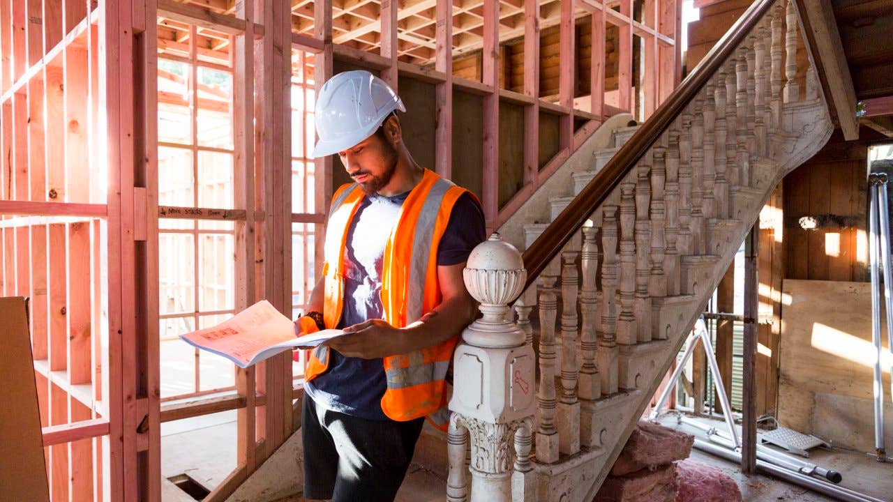 A builder sits on the stairs inside a house under renovation and studies the building plans