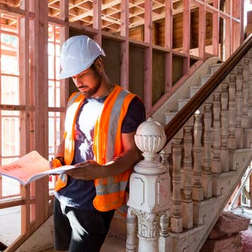 A builder sits on the stairs inside a house under renovation and studies the building plans