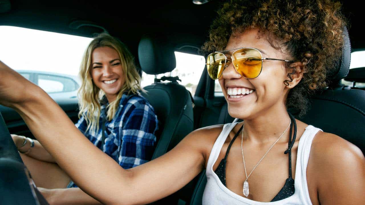 Portrait of two women with long blond and brown curly hair sitting in car, wearing sunglasses, smiling.
