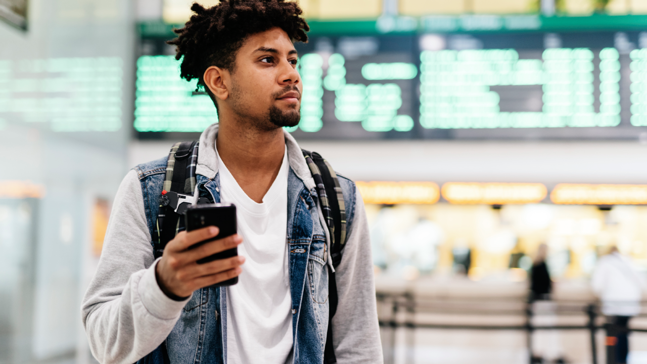 Young man inside the airport holding a mobile phone.young afro man holding a mobile phone looking at the departures of planes in an airport