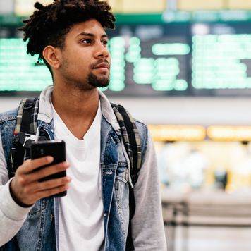 Young man inside the airport holding a mobile phone.young afro man holding a mobile phone looking at the departures of planes in an airport