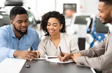 Spouses Signing Papers With Auto Seller In Dealership Office