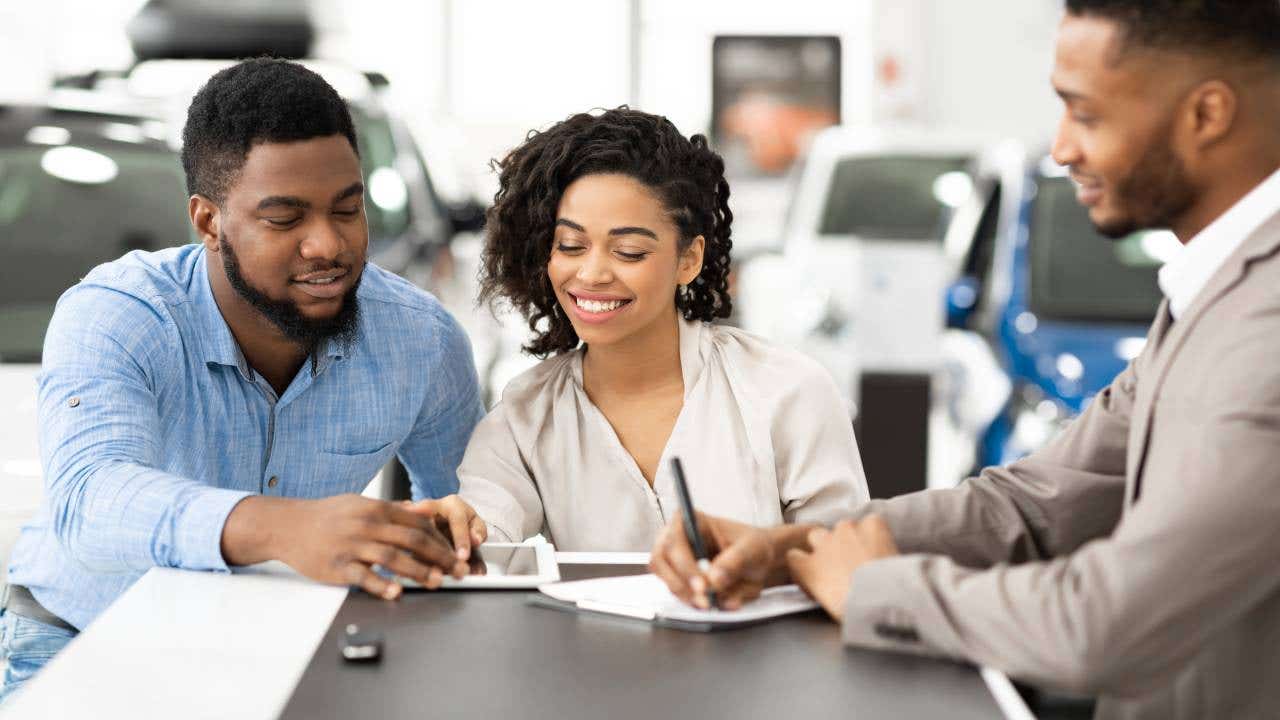 Spouses Signing Papers With Auto Seller In Dealership Office