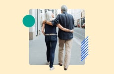 An old couple walking down the sidewalk with their arms around each other's shoulders.