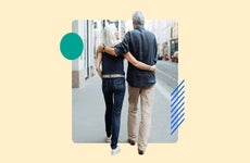 An old couple walking down the sidewalk with their arms around each other's shoulders.