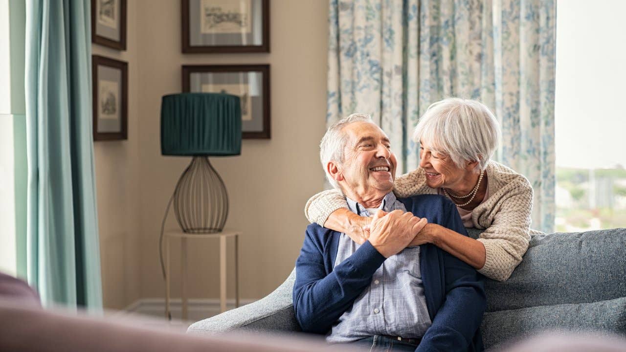 Happy senior woman embracing her husband at home while laughing together.