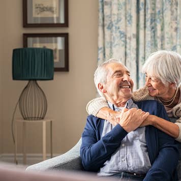 Happy senior woman embracing her husband at home while laughing together.