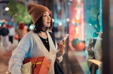 Beautiful young Asian woman holding smart phone and Christmas gifts, looking at shop window on shopping street. Christmas shopping. Online shopping. Contactless payment.