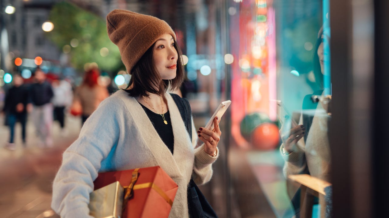 Beautiful young Asian woman holding smart phone and Christmas gifts, looking at shop window on shopping street. Christmas shopping. Online shopping. Contactless payment.