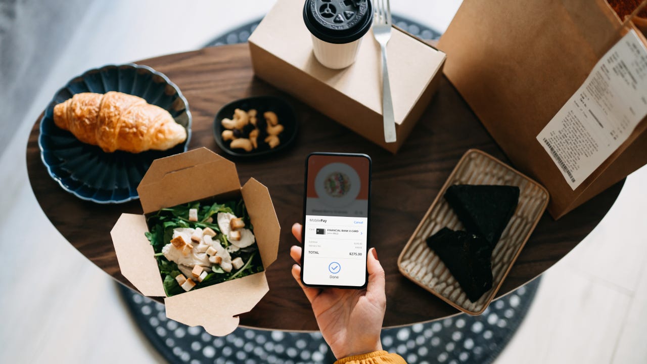 Flat lay of woman's hand holding smartphone with takeaway food ordering mobile app, making mobile payment with credit card, with assorted takeaway dishes served on coffee table. Eating at home and technology concept