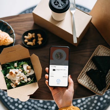 Flat lay of woman's hand holding smartphone with takeaway food ordering mobile app, making mobile payment with credit card, with assorted takeaway dishes served on coffee table. Eating at home and technology concept