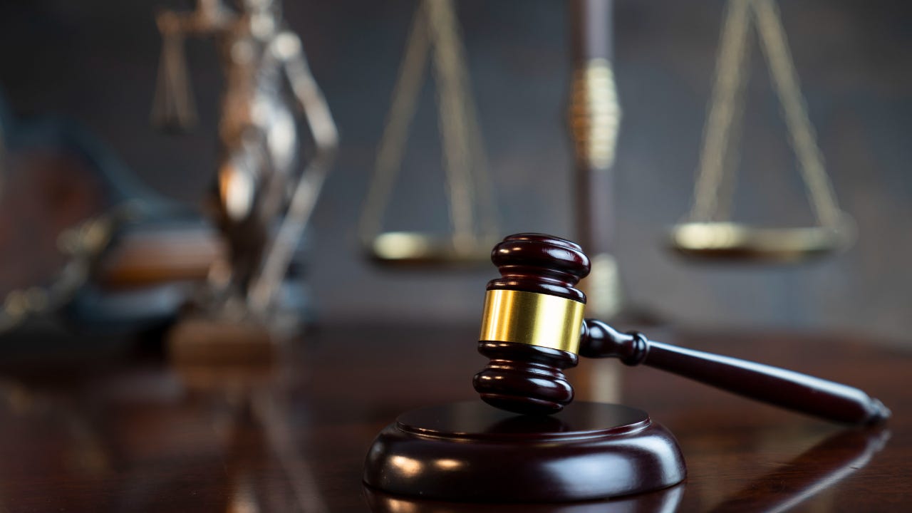 A gavel rests on a wooden table. Out of focus in the background are the scales of justice.