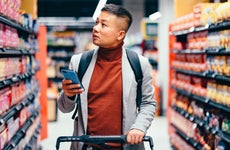 Handsome Asian male walking down the product aisle in the supermarket, looking at shelves and searching for groceries from the list on his mobile phone he is holding in his hand.