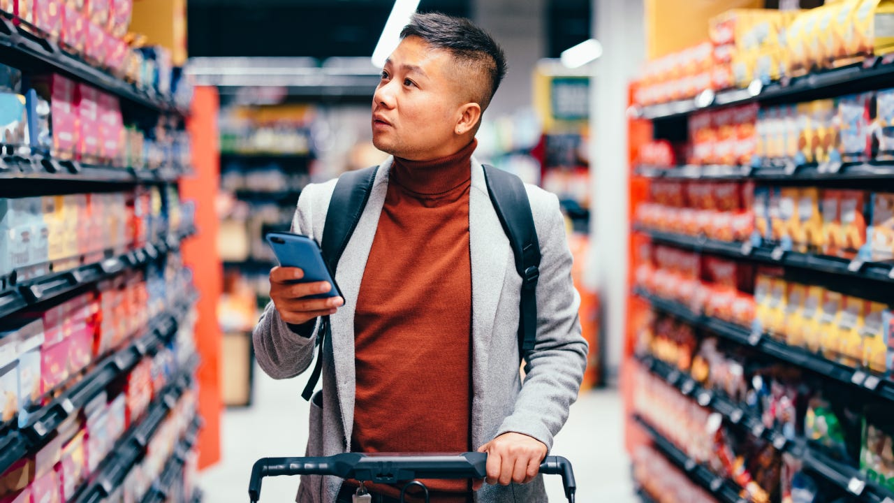 Handsome Asian male walking down the product aisle in the supermarket, looking at shelves and searching for groceries from the list on his mobile phone he is holding in his hand.