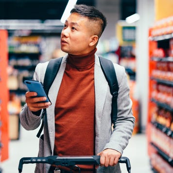 Handsome Asian male walking down the product aisle in the supermarket, looking at shelves and searching for groceries from the list on his mobile phone he is holding in his hand.