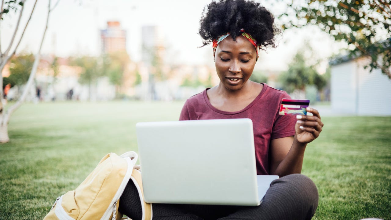 Portrait of a beautiful young African American woman sitting on the grass in the park and shopping online.
