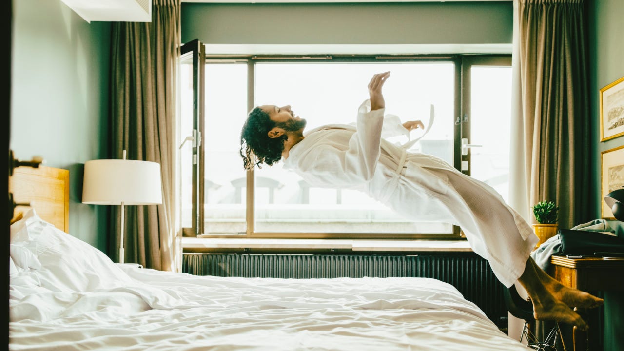 Playful man wearing robe jumping on bed at hotel