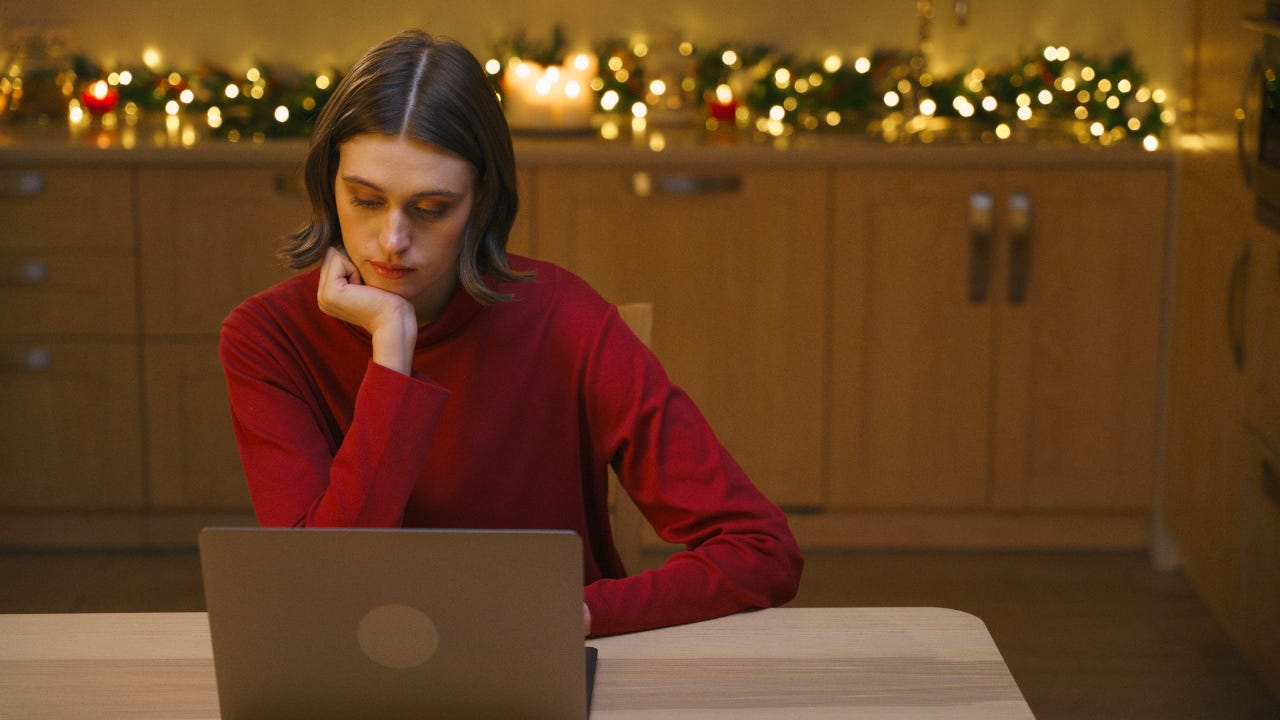 Person in red long-sleeve shirt sitting at laptop looking thoughtful with winter holiday decorations in the background