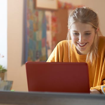 Woman in an orange shirt sitting at table while smiling at her laptop.