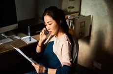 Serious mid adult Asian woman listening on phone, sitting in office chair in dark room, holding paperwork, checking accounts, financial planning, money concerns, difficulty, debt