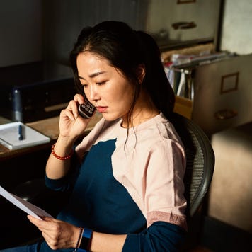 Serious mid adult Asian woman listening on phone, sitting in office chair in dark room, holding paperwork, checking accounts, financial planning, money concerns, difficulty, debt