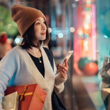 Beautiful young Asian woman holding smart phone and Christmas gifts, looking at shop window on shopping street. Christmas shopping. Online shopping. Contactless payment.