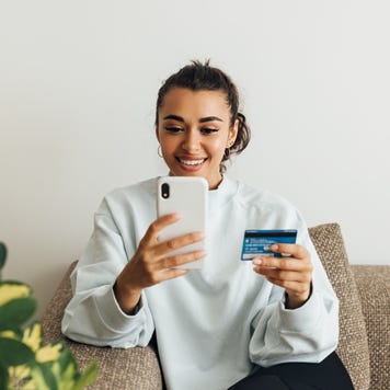 Smiling Woman Holding Credit Card While Using Smart Phone At Home - stock photo
