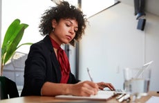 Confident young female financial advisor writing on diary while sitting with laptop at desk in office