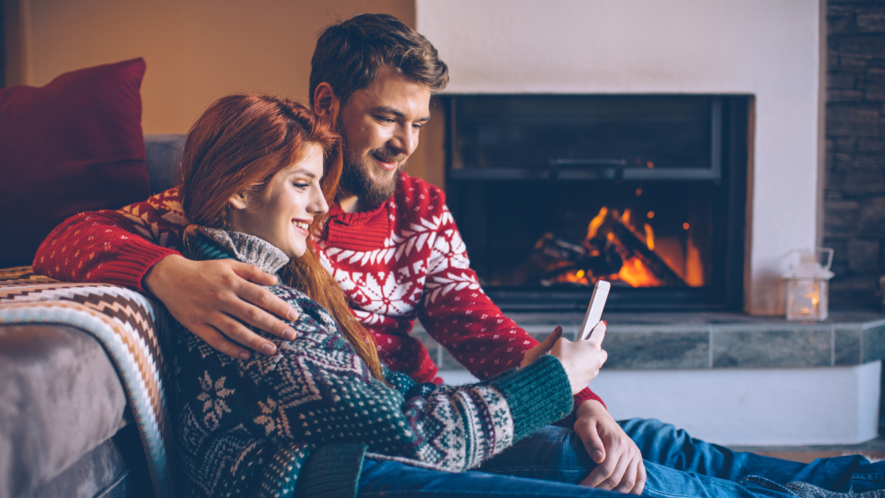 Couple on vacation at mountain cabin. Checking smart phone for news or messages. Sitting on the floor on a fur by fireplace in cosy living room on Christmas. Wearing festive knitted sweaters and socks.