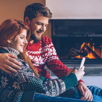 Couple on vacation at mountain cabin. Checking smart phone for news or messages. Sitting on the floor on a fur by fireplace in cosy living room on Christmas. Wearing festive knitted sweaters and socks.