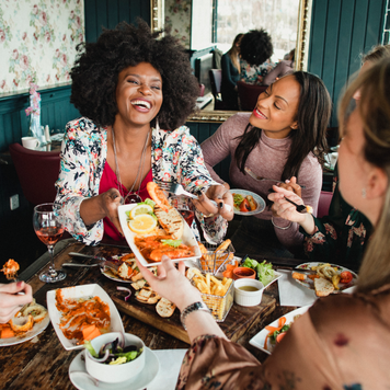 A group of women are enjoying a meal in a restaurant with rose wine. They have a king prawns sharing platter , one woman can be seen passing her friend a plate of food. Mixed ethnic group