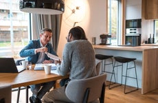 Real estate agent and young couple sitting around a table and discussing about the price and blueprints of a new residential building project. They are indoors in a new apartment.