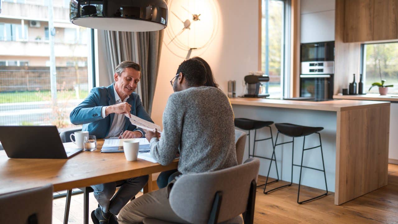 Real estate agent and young couple sitting around a table and discussing about the price and blueprints of a new residential building project. They are indoors in a new apartment.