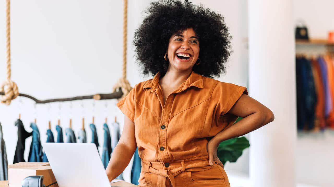 A Black female entrepreneur with natural hair smiles in front of a clothing rack.