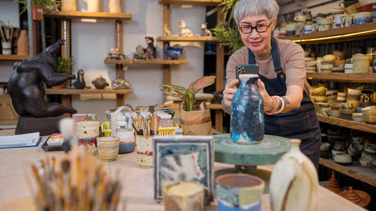 An older Asian woman in a pottery studio photographs a vase.